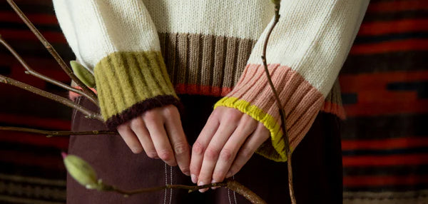 Person wearing a striped sweater with green, brown, and pink cuffs holding branches.