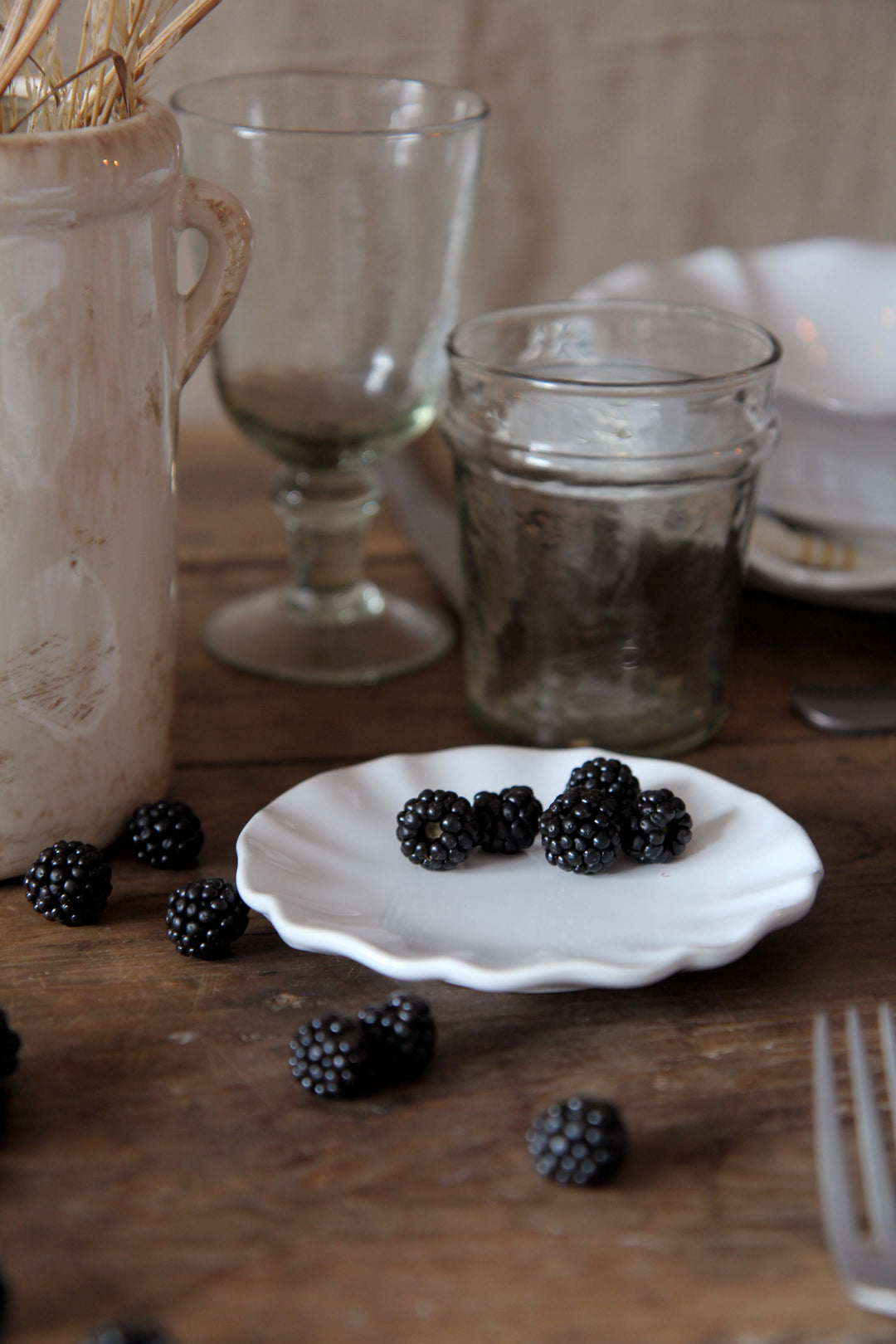 Blackberries on a white plate with glasses and a vase in the background on a wooden table.