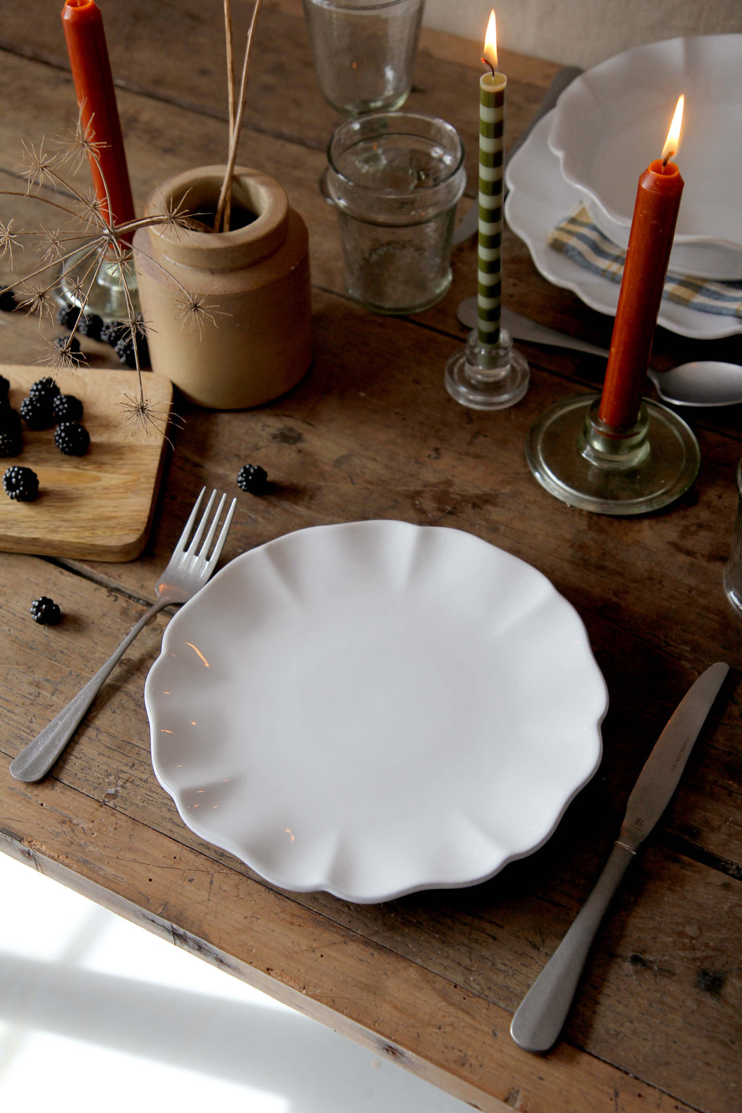 Dining table setting with white scalloped plates, candles, and cutlery on a wooden surface.