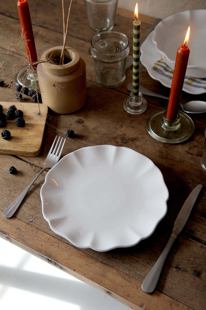 Dining table setting with white scalloped plates, candles, and cutlery on a wooden surface.