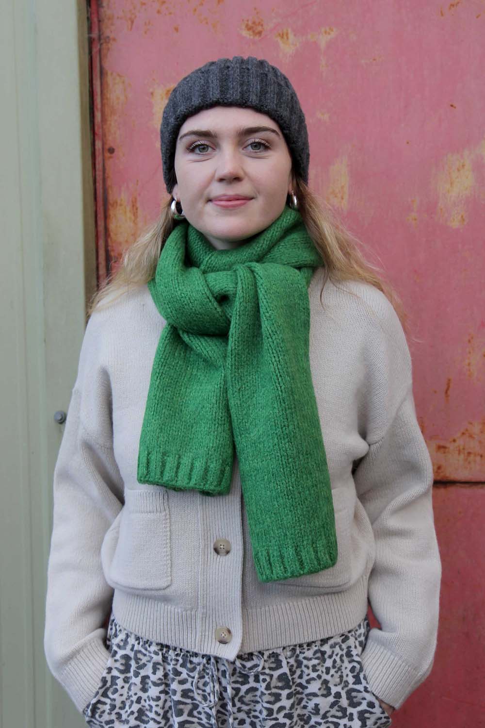 model wearing green woollen scarf and grey beanie against a pink backdrop