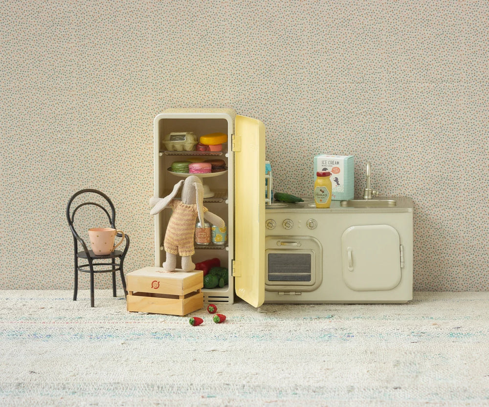Miniature kitchen set with refrigerator, oven, and chair against a textured wall.