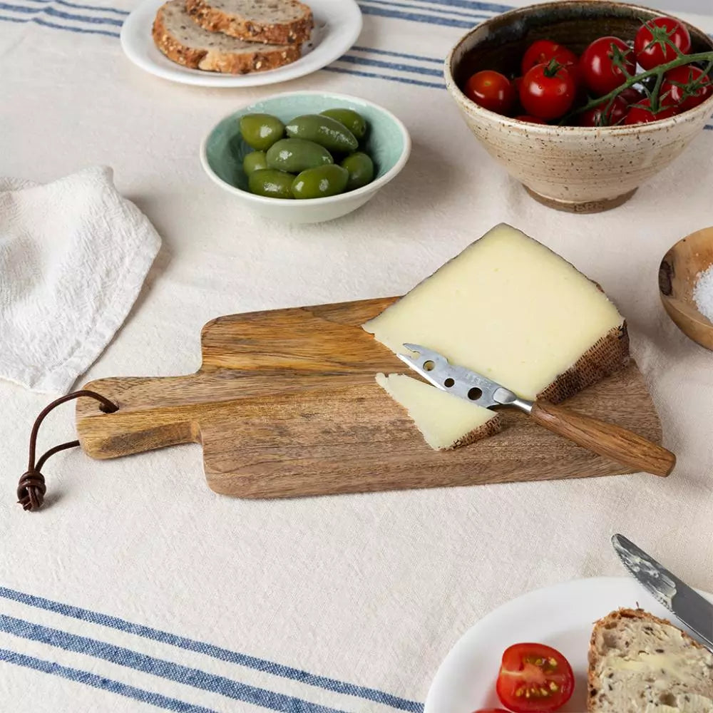 Wooden cheese board with a wedge of cheese, olive, and tomato on a tablecloth.