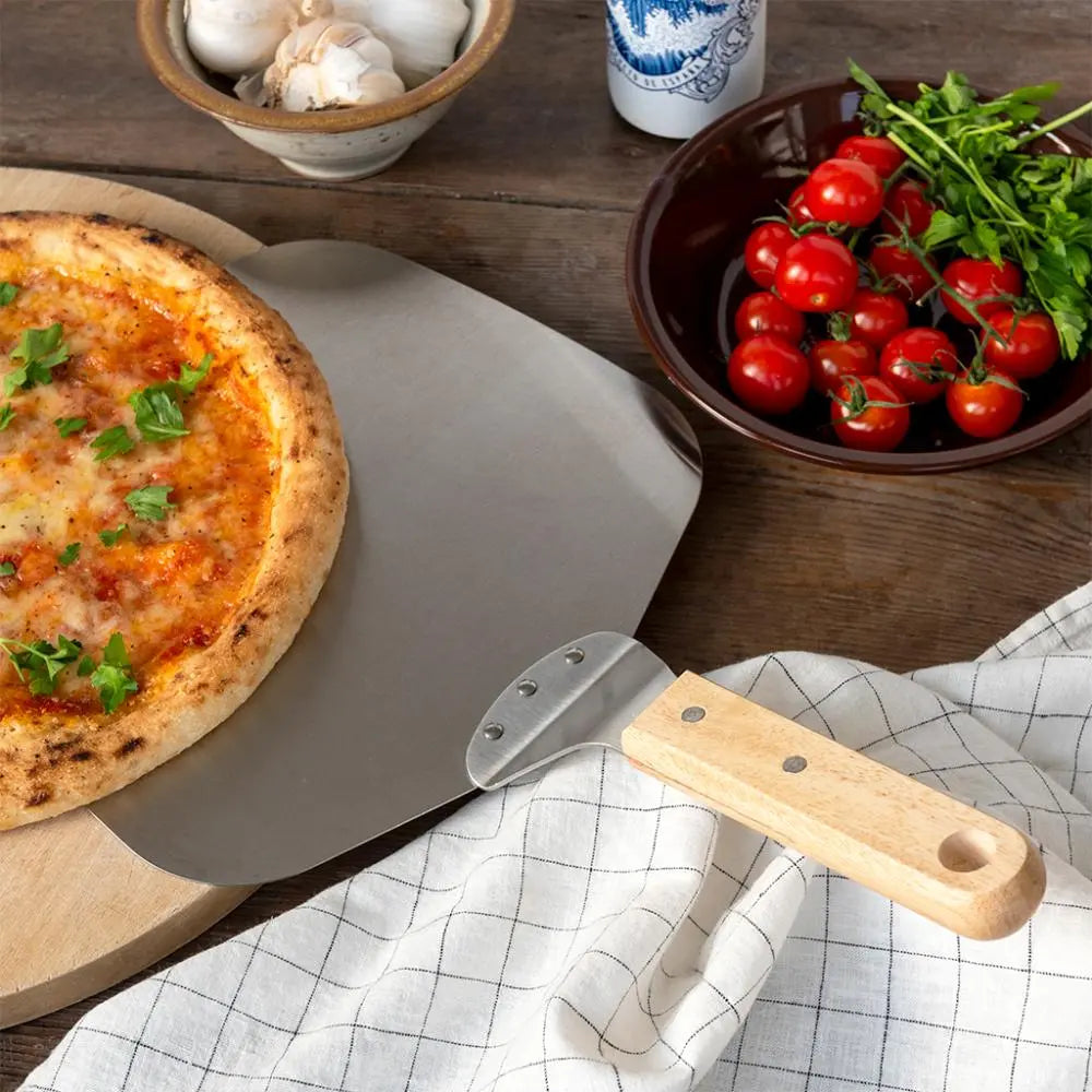 Pizza on a wooden board with a pizza peel, bowl of cherry tomatoes, and garlic on a wooden table.