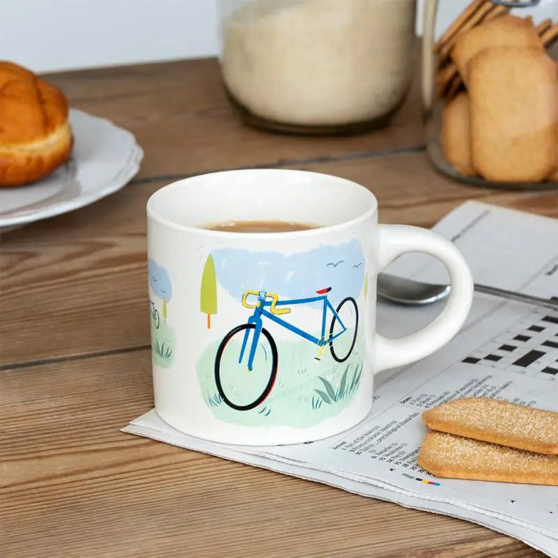 White mug with bicycle design on a wooden table with coffee, cookies, and newspaper.