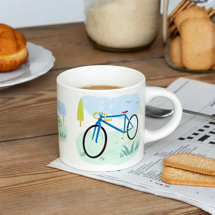 White mug with bicycle design on a wooden table with coffee, cookies, and newspaper.