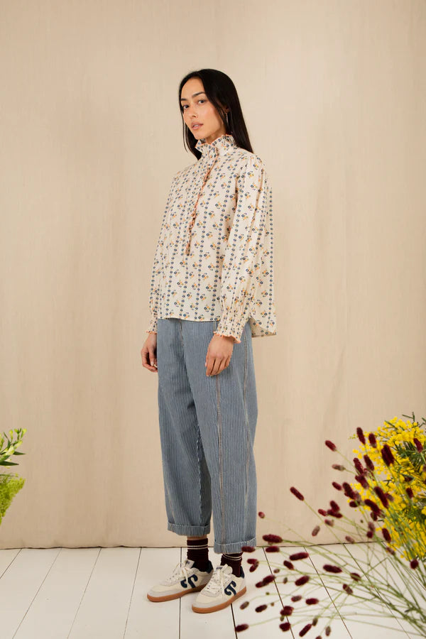 Woman wearing a patterned blouse and blue jeans standing against a beige wall with floral decorations.