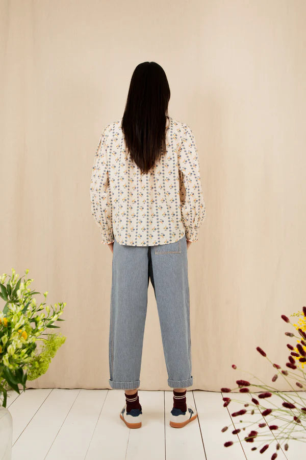 Person wearing a patterned blouse and jeans standing against a beige wall with flowers on either side.