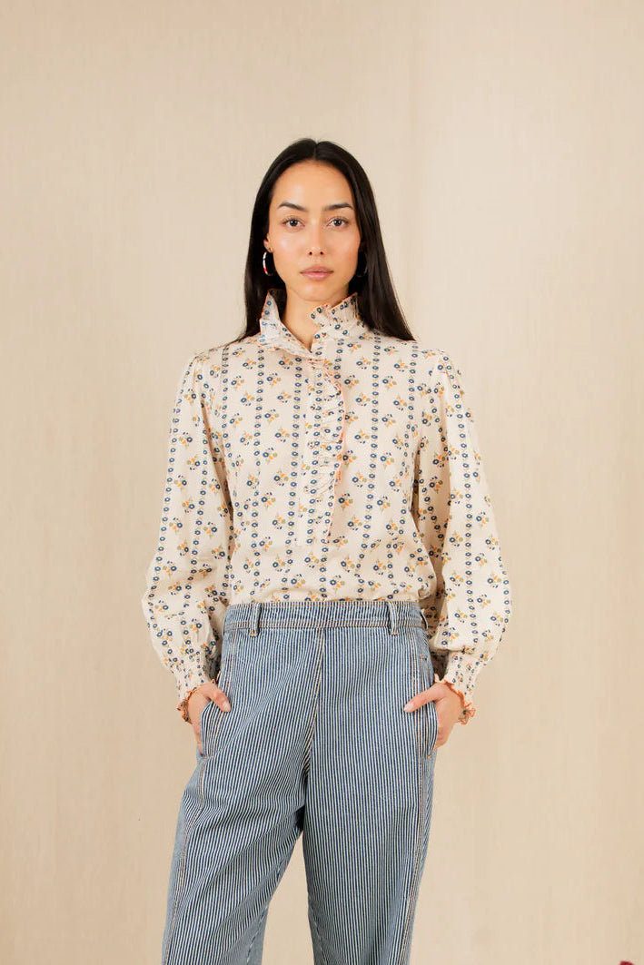 Woman wearing a patterned blouse and blue jeans standing against a beige wall with flowers on the floor.