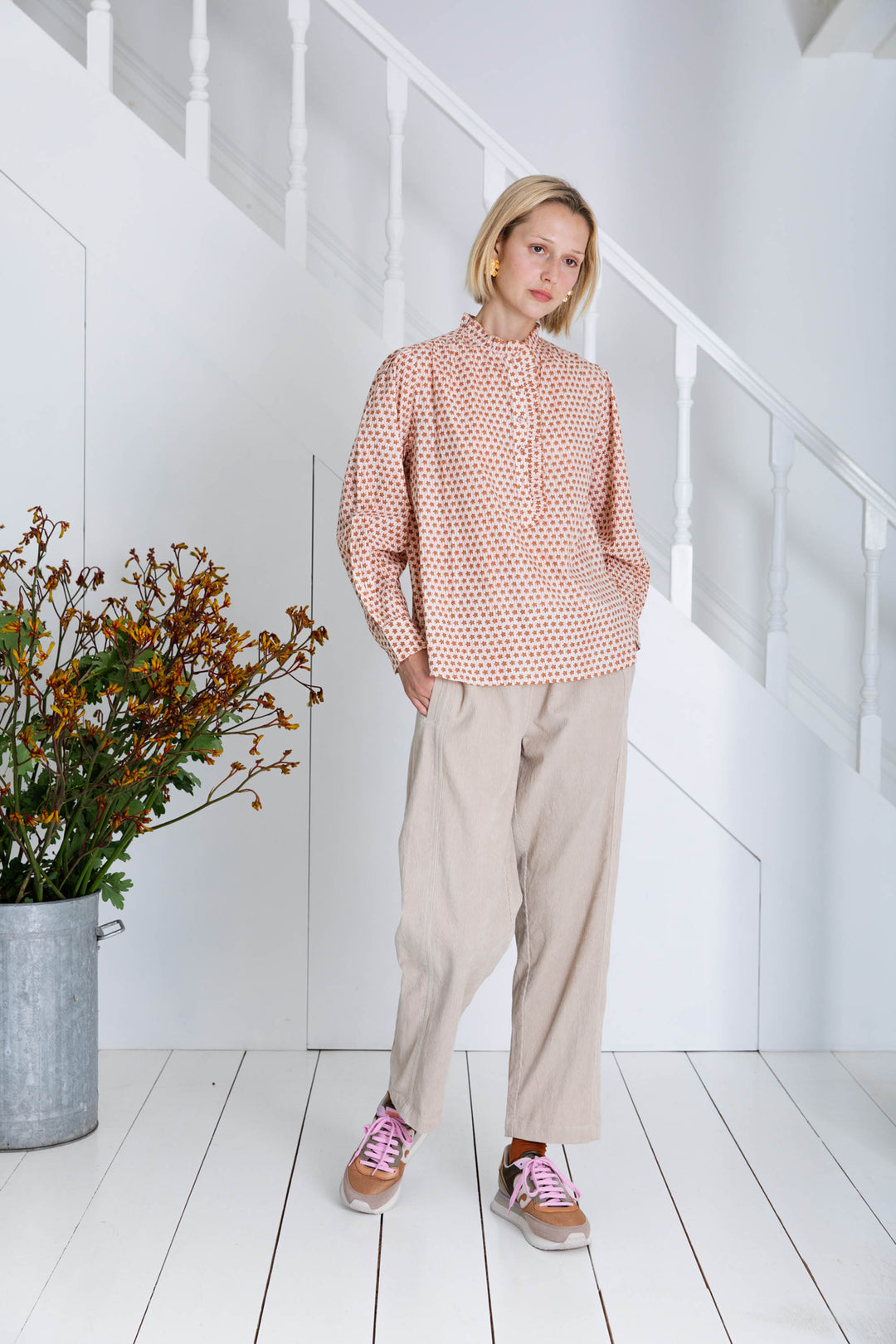 Woman wearing a pink patterned blouse and beige pants standing in a minimalistic room with white walls and wooden floor.