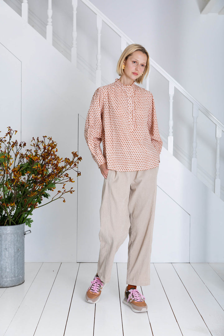 Woman wearing a pink patterned blouse and beige pants standing in a minimalistic room with white walls and wooden floor.