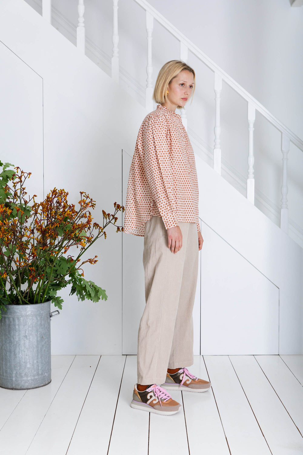 Woman standing in a minimalistic room with white walls and wooden floor, wearing a patterned shirt and beige pants.