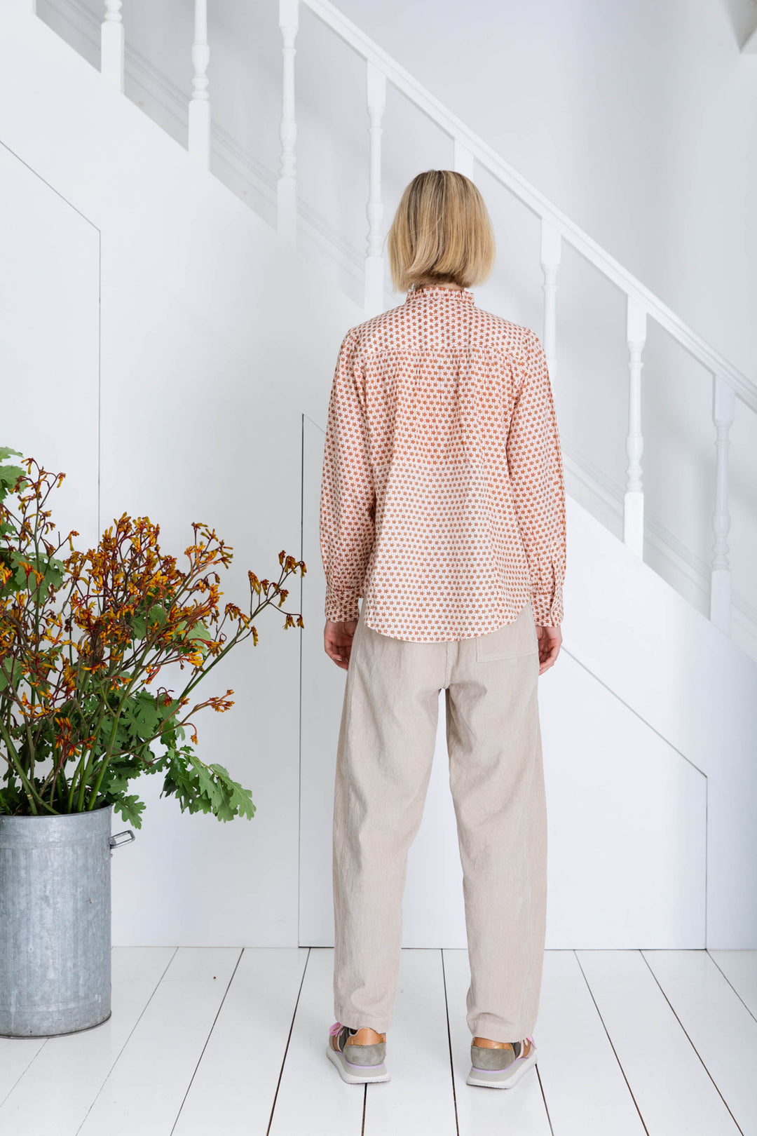 Person wearing a patterned shirt and beige pants standing in a room with white walls and wooden floor.