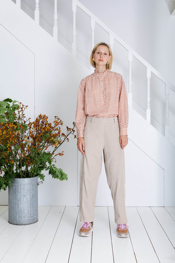 Woman in a polka dot blouse and beige pants standing in a room with white walls and wooden floor.
