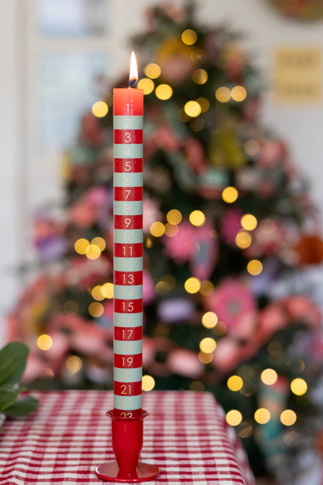 A red and white striped advent candle with numbered sections from 1 to 21 stands lit on a red-and-white checkered tablecloth in front of a blurred Christmas tree with warm lights.
