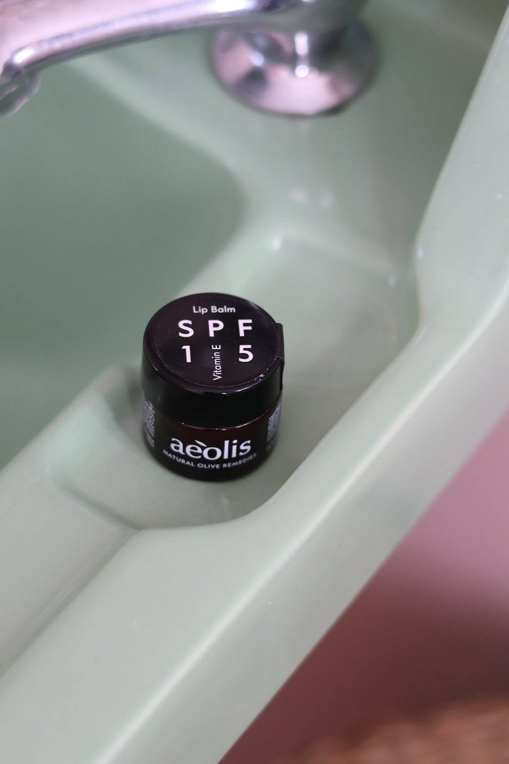 A small jar of aeolis Lip Balm SPF 15 in shade E, placed inside a pale green ceramic sink basin.
