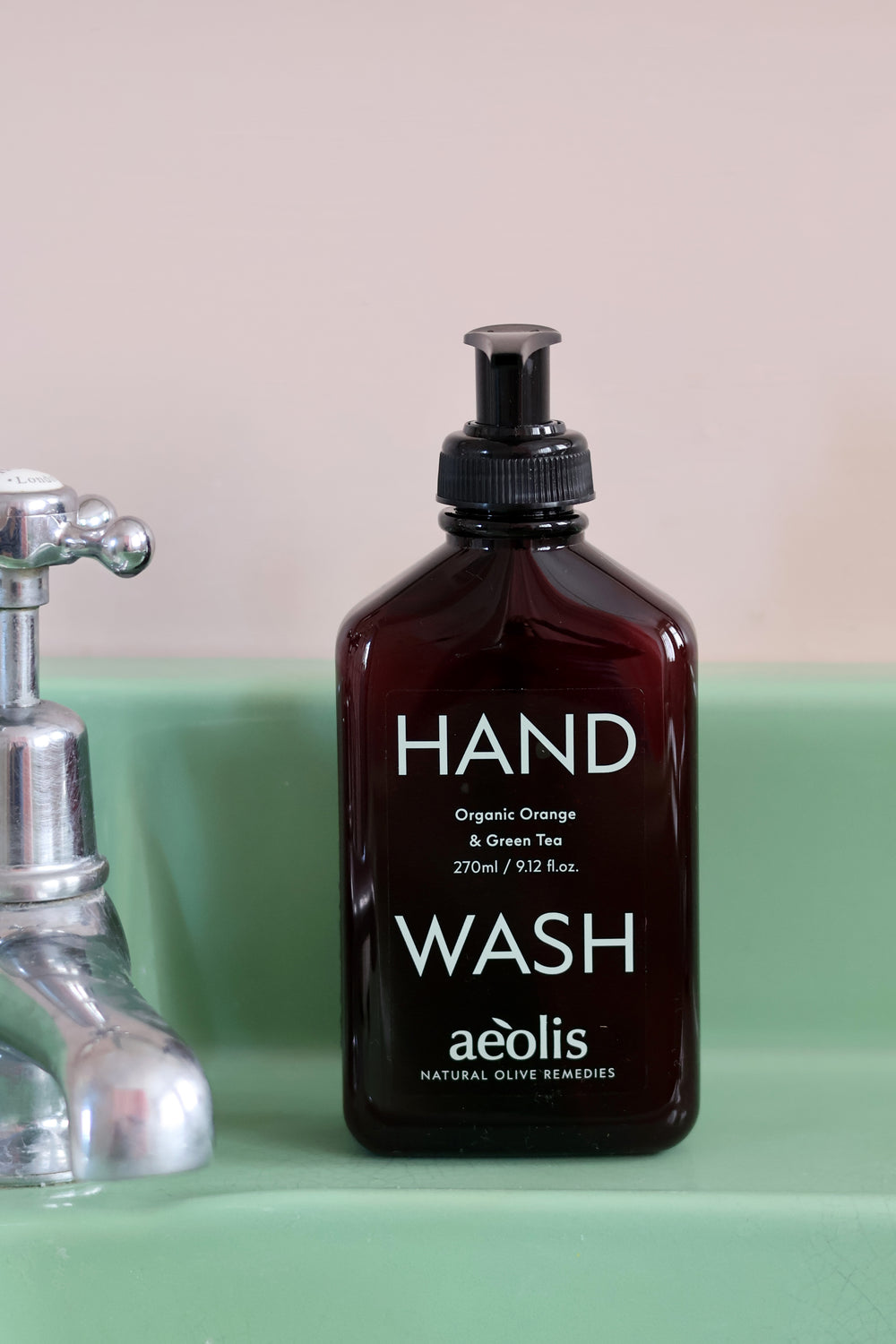 A bottle of aedis hand wash in a dark amber glass bottle with a black pump dispenser, set on a mint green surface next to a chrome faucet.