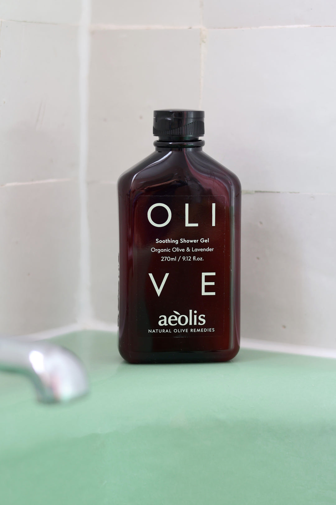 A bottle of aéolis OLI VE Soothing Shower Gel standing on a mint green surface against a white tiled wall.