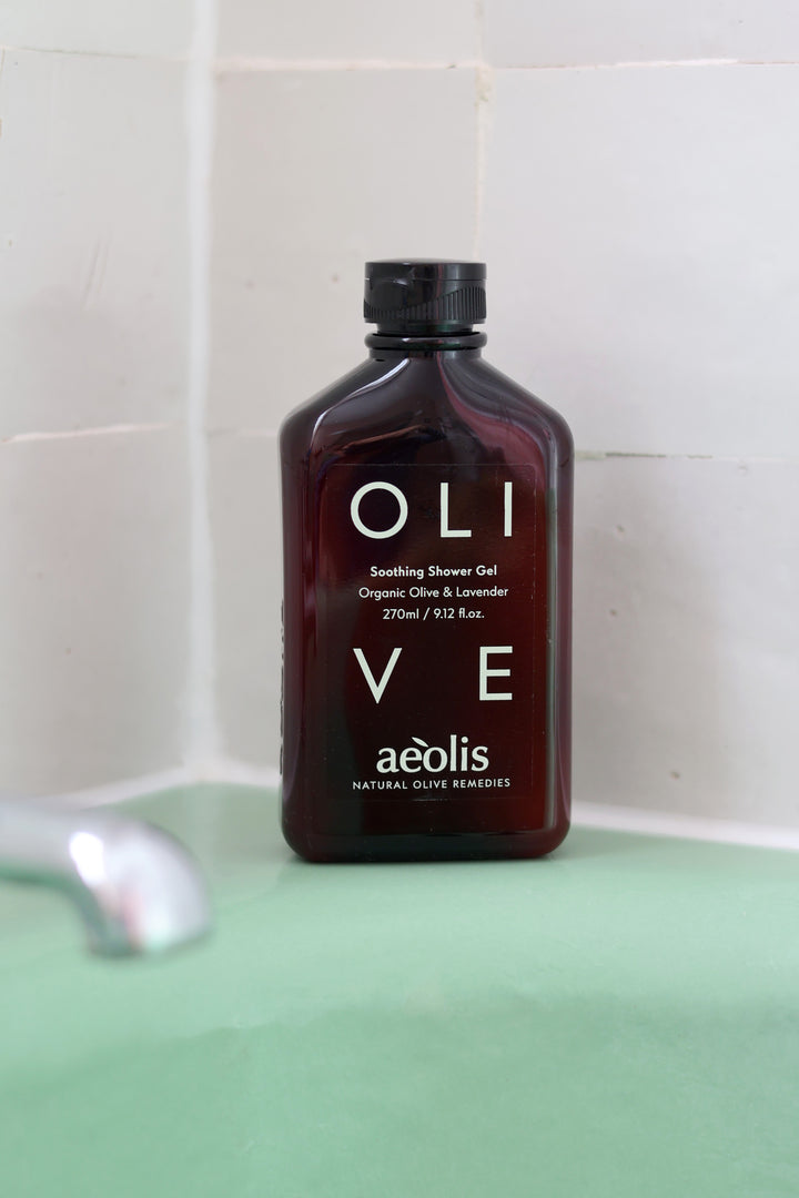 A bottle of aéolis OLI VE Soothing Shower Gel standing on a mint green surface against a white tiled wall.