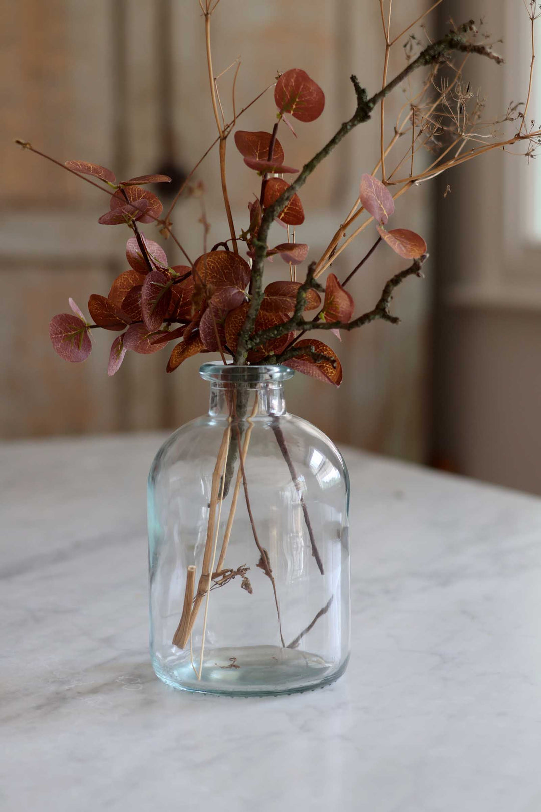 A clear glass bottle vase filled with dried botanical stems and round reddish-brown leaves, placed on a light marble surface with a softly blurred indoor background.