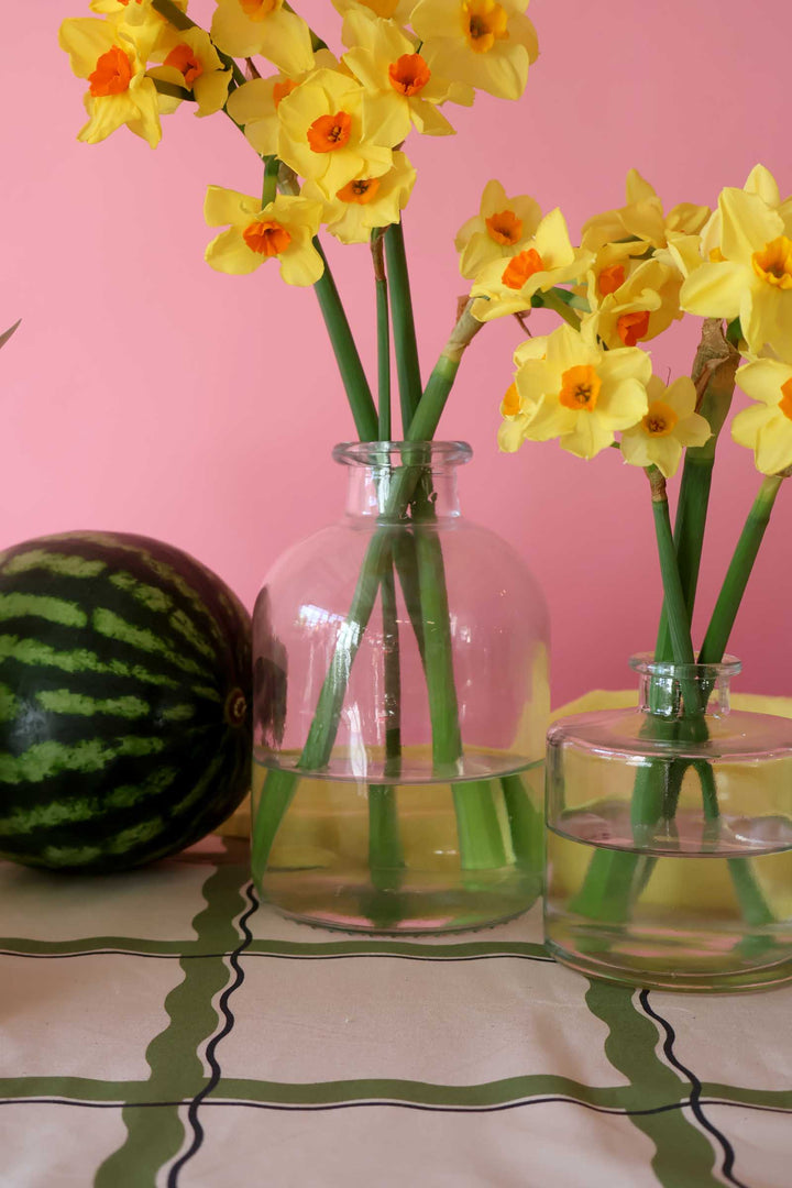 Two glass vases with yellow flowers and a watermelon on a patterned tablecloth against a pink wall.