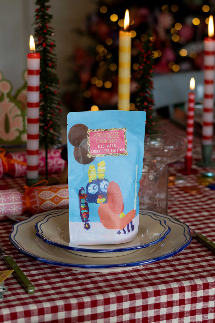 A stand-up pouch of chocolate milk powder with a light blue background and cartoon illustrations, displayed on a plate on a red-and-white checkered tablecloth with candles and holiday decor in the background.