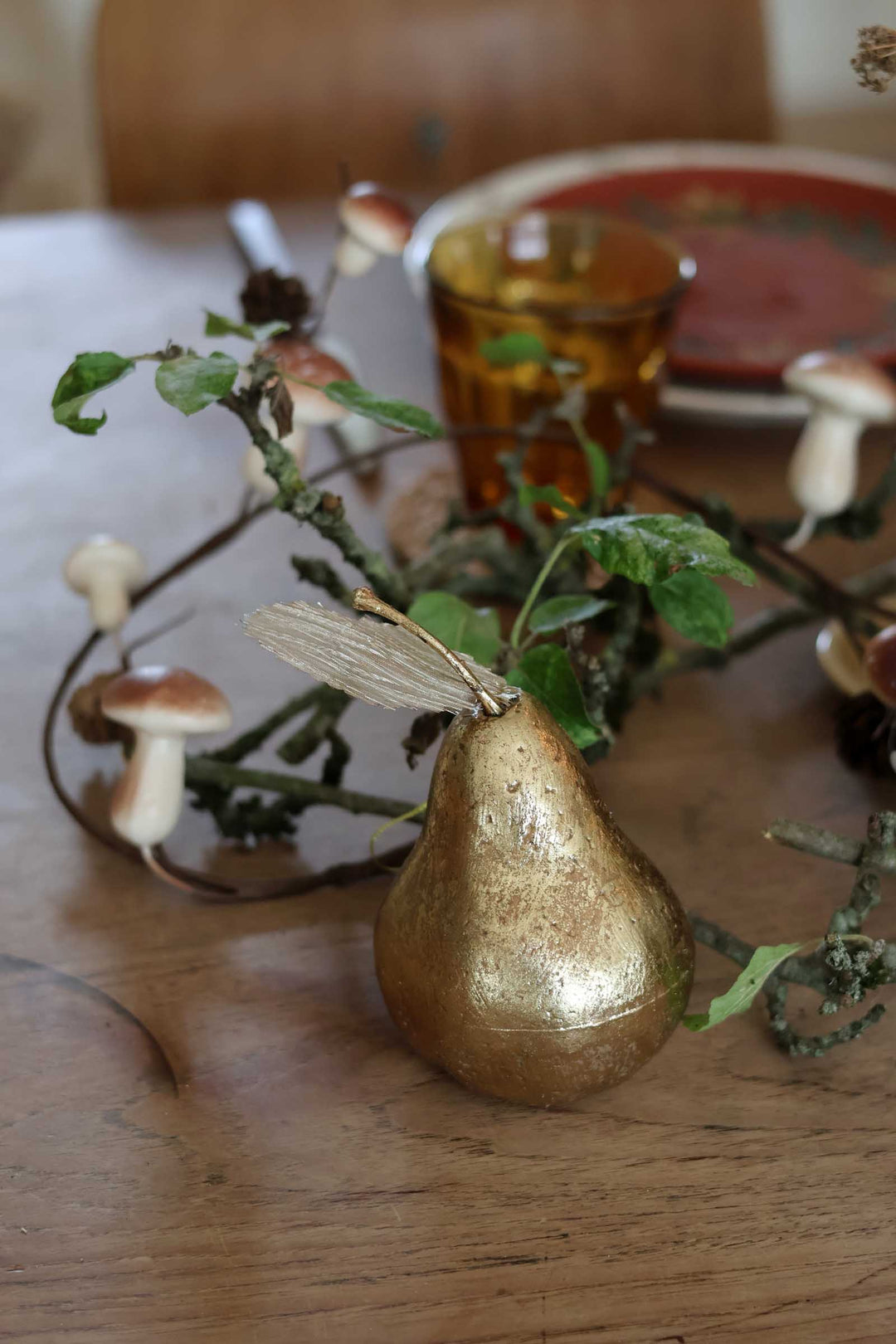 A rustic tabletop scene with a metallic gold pear-shaped centerpiece, green leaves, small mushrooms, and a blurred amber glass in the background.