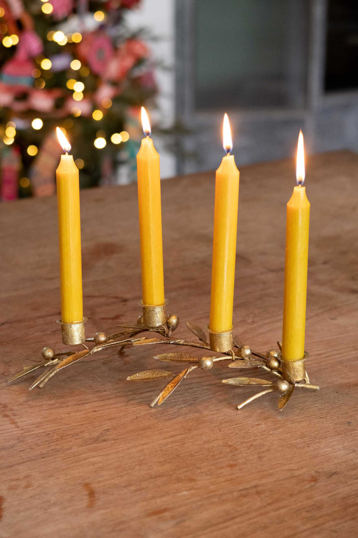 Four lit yellow taper candles set in a gold-toned metal candelabra with leaf and berry details, placed on a wooden tabletop with a softly blurred holiday background.