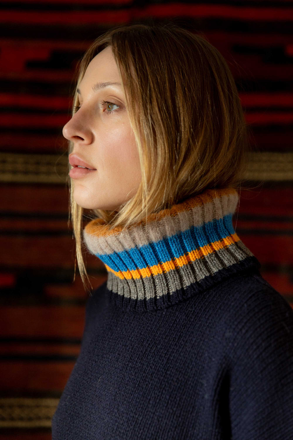 Woman wearing a dark blue sweater with a colorful striped turtleneck against a red brick wall.