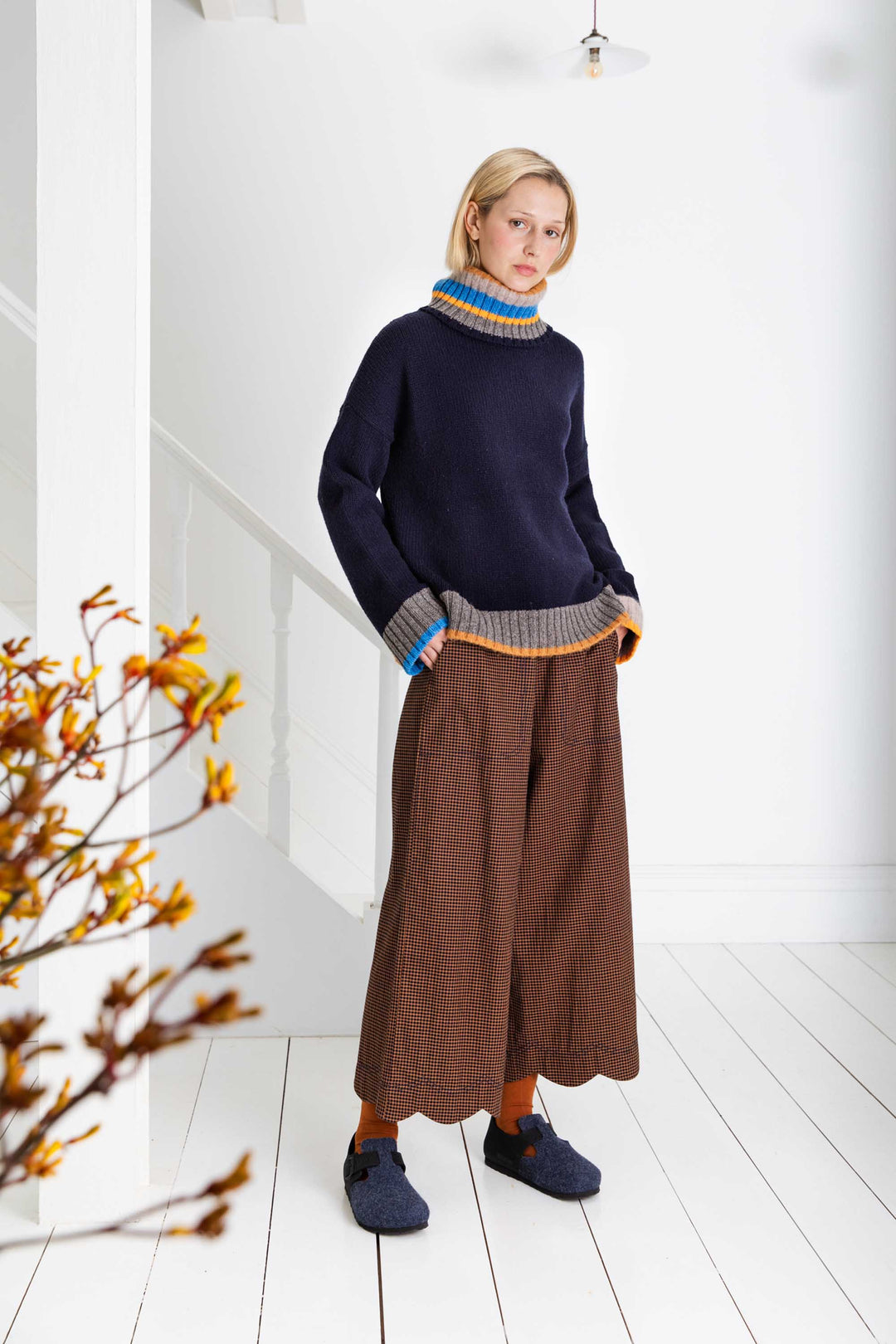 Woman wearing a dark sweater with colorful trim and brown wide-leg pants in a minimalistic setting.