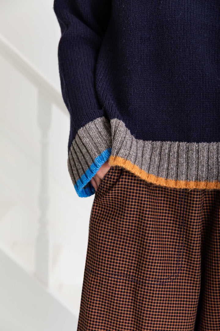 Navy sweater with multicolored cuffs and hem paired with brown checkered pants on a white background