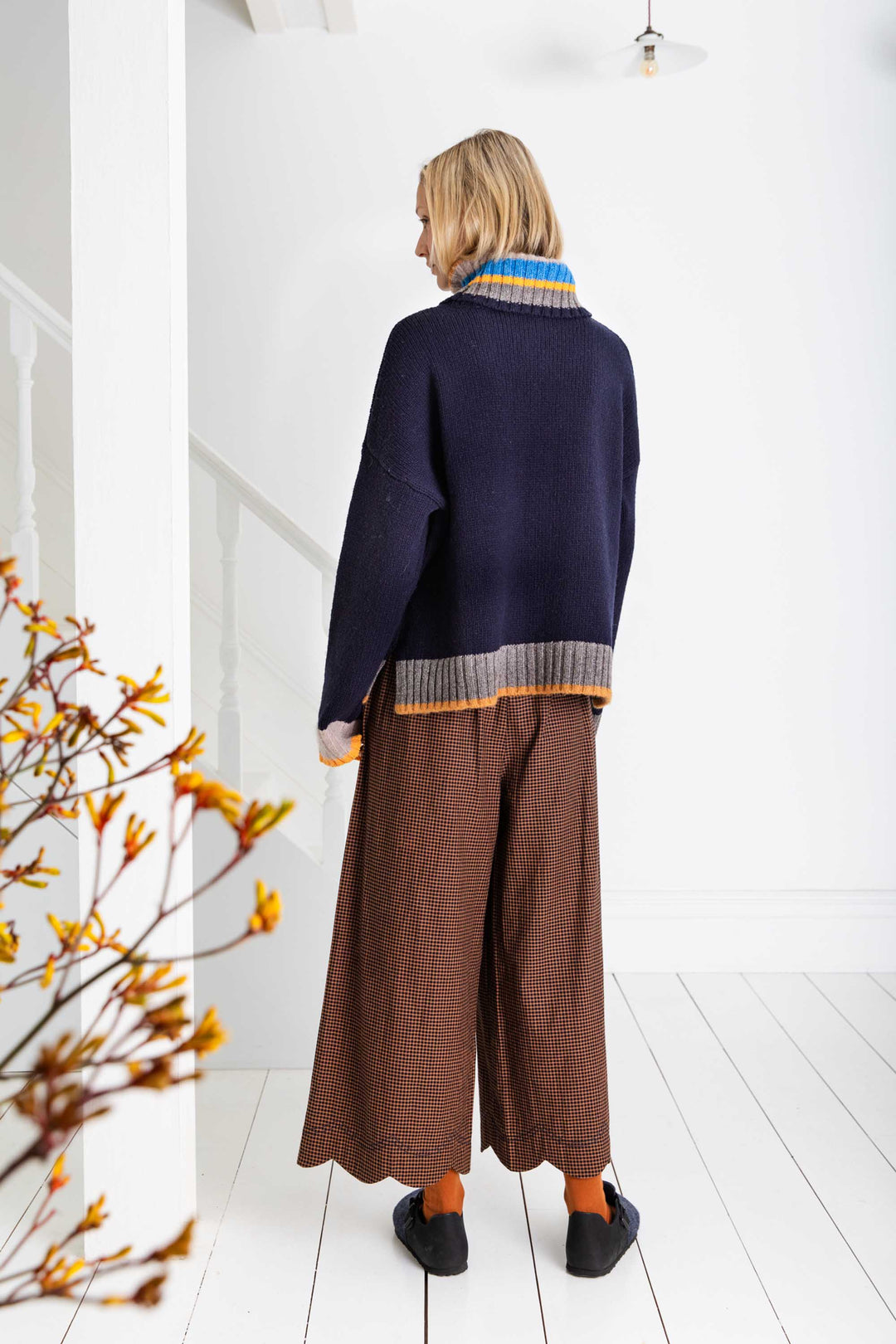 Person wearing a navy sweater with a colorful collar and brown wide-leg pants in a minimalistic room.