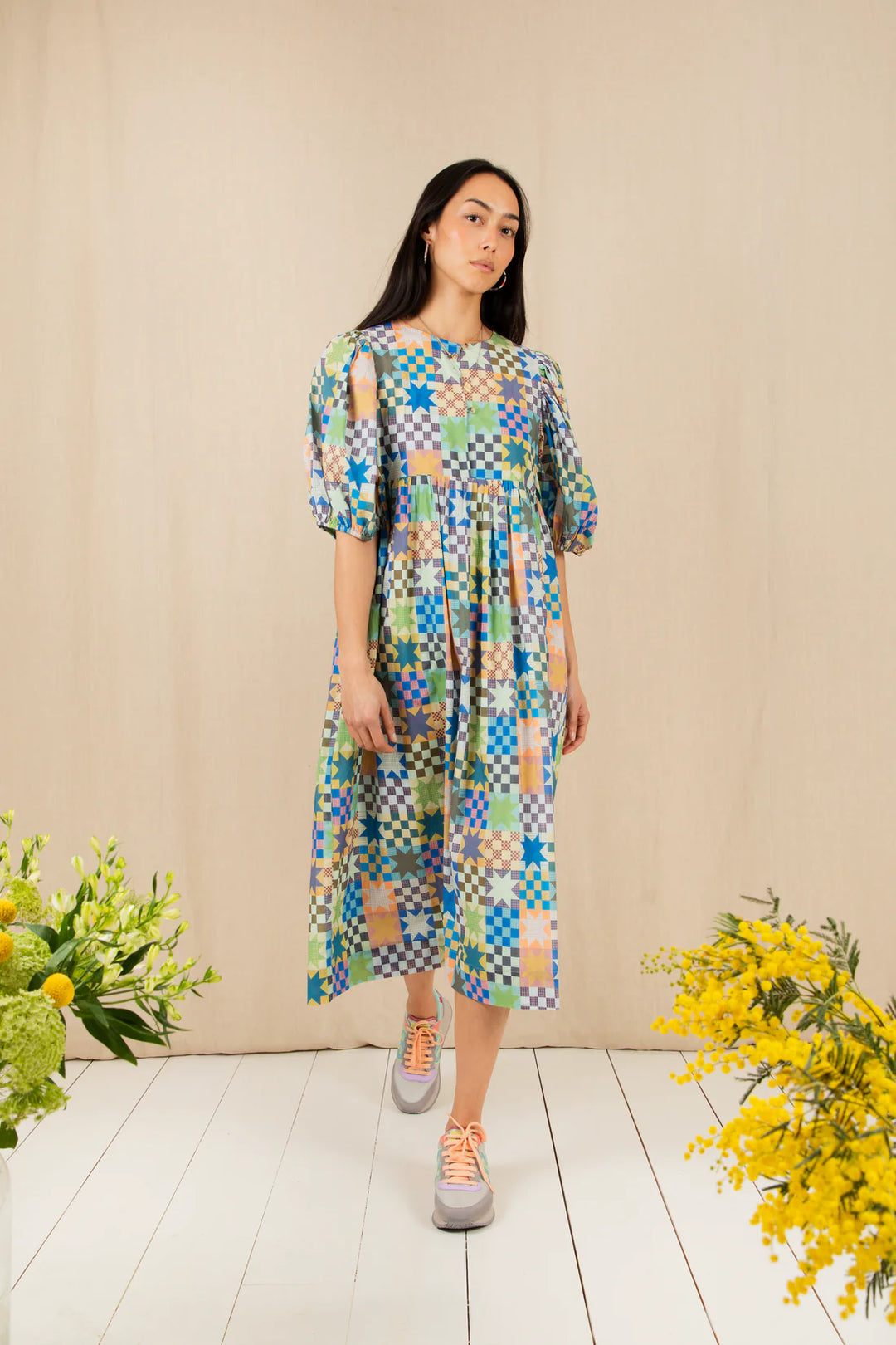 Woman wearing a colorful patterned dress standing against a beige wall with flowers on either side.