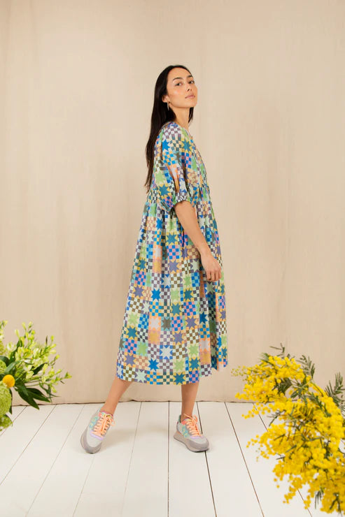 Woman wearing a colorful patterned dress standing against a beige wall with flowers on either side.