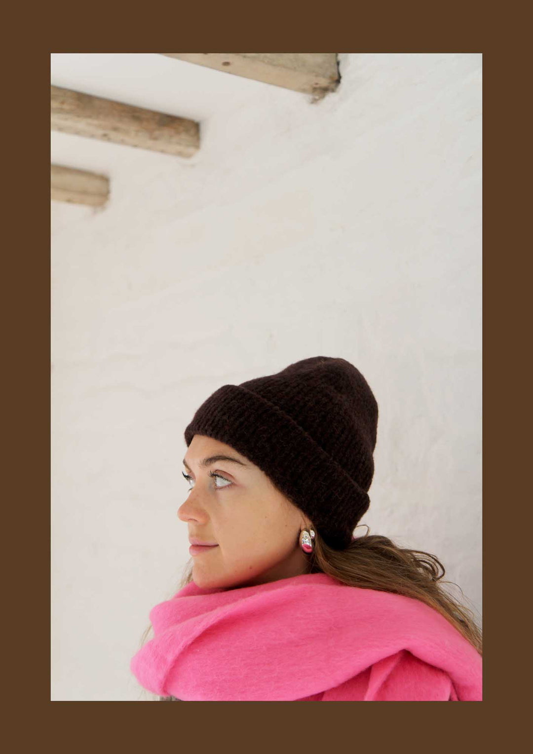 Woman wearing a brown knit beanie and pink scarf against a white wall.