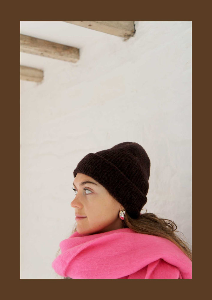 Woman wearing a brown knit beanie and pink scarf against a white wall.