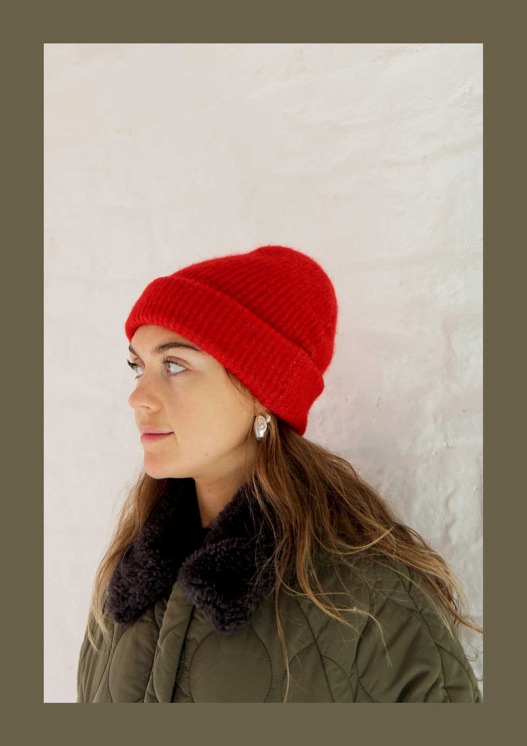 Person wearing a red knit beanie and green coat with fur collar against a light background