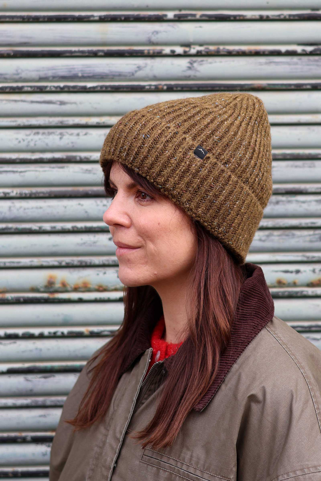 A woman in profile wearing a brown ribbed knit beanie hat and a brown jacket, standing in front of a weathered metal shutter.