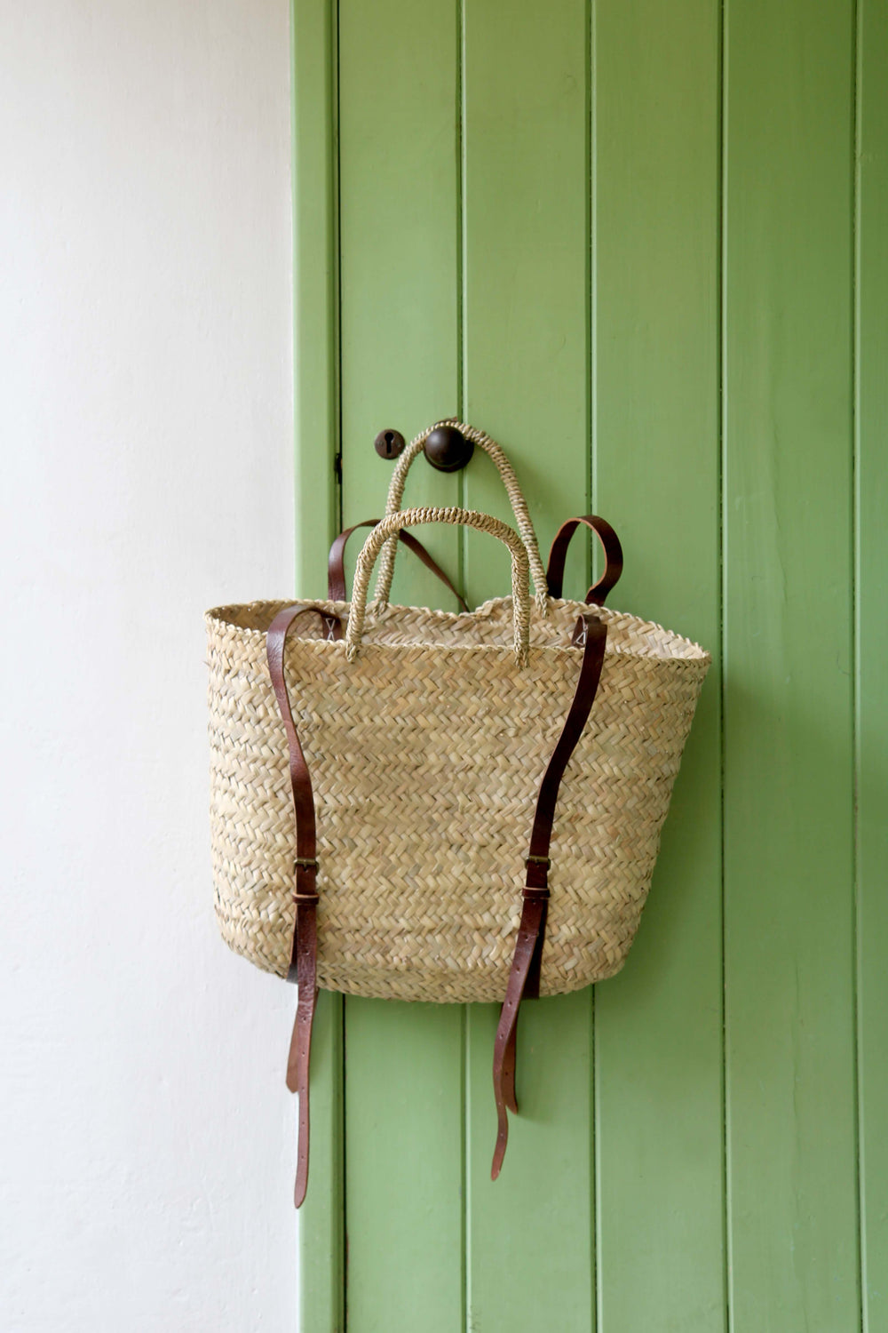 Woven bag with brown straps hanging on a green door