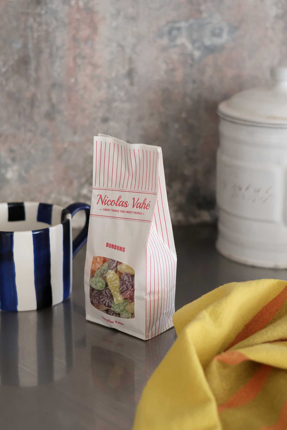 A bag of Nicolas Vache Bresseau cheese sticks stands on a countertop in a kitchen setting, with a blue and white striped mug on the left and a yellow cloth in the foreground.