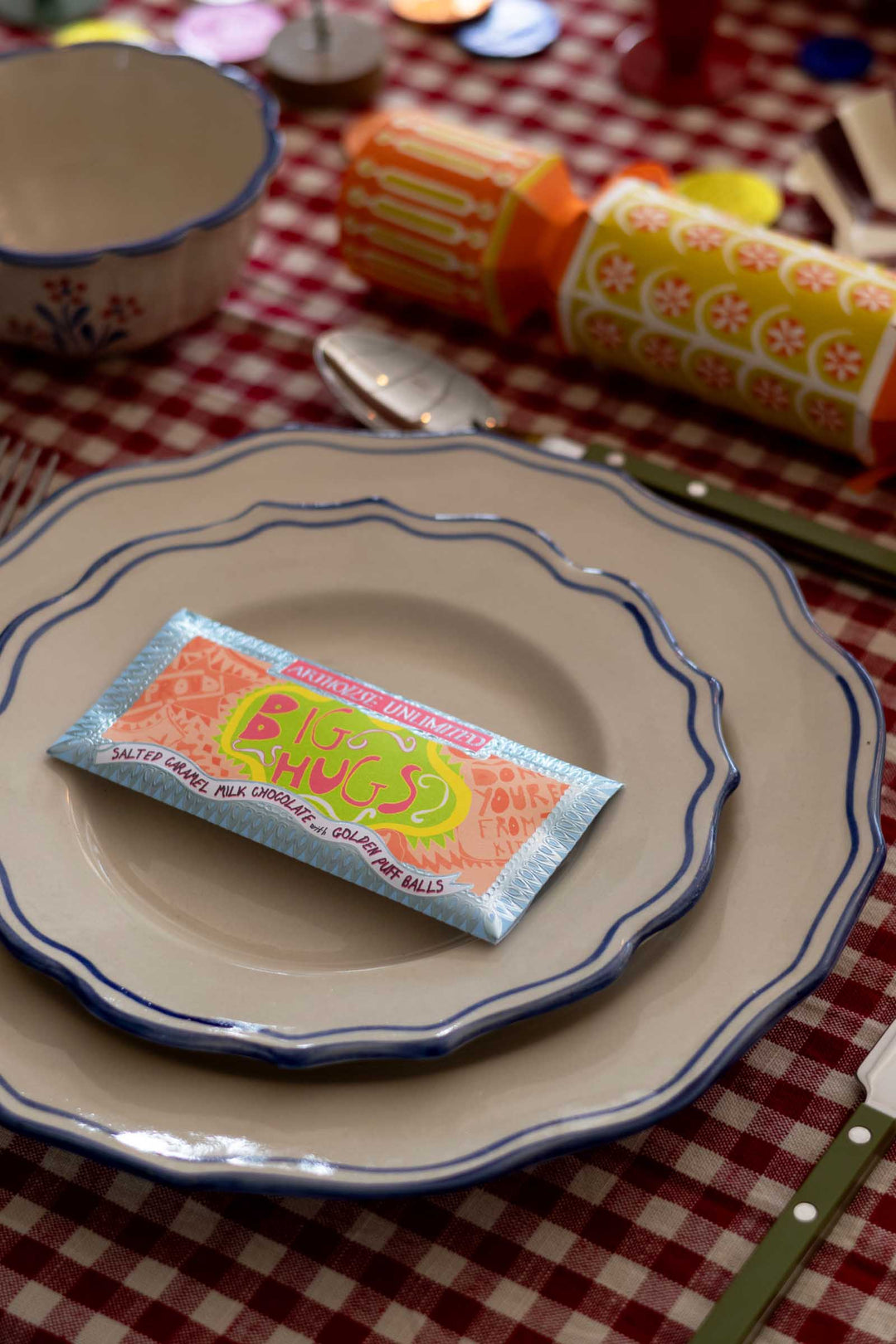 A packet of Cluck Cluck Candy salted caramel tea cookies and poppy seed balls rests on a scalloped cream plate with blue trim, set on a red and white checkered tablecloth with cutlery and other dishes blurred in the background.