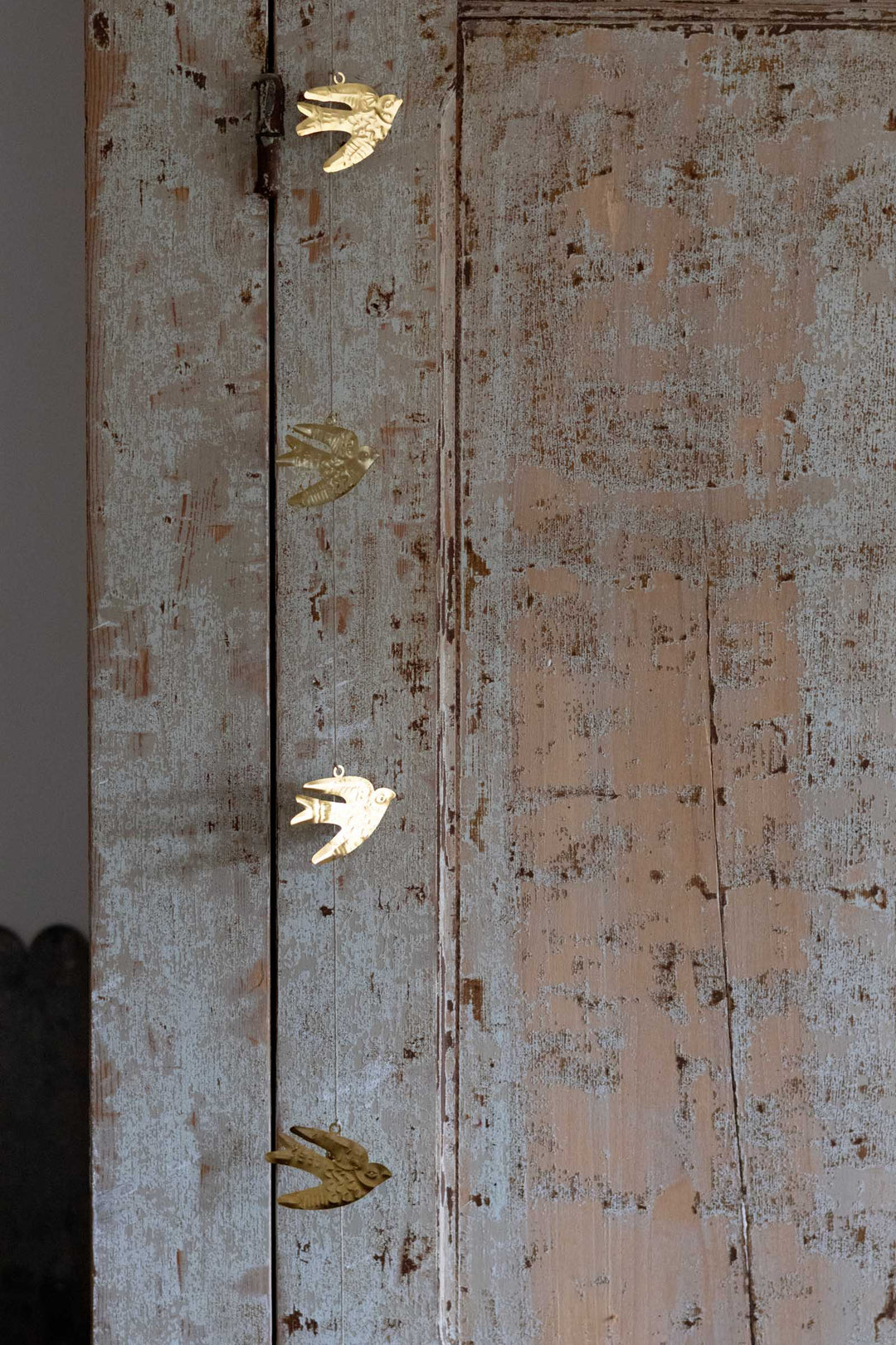 A close-up of a distressed, weathered wooden door with a light gray and peach-pink peeling paint finish, decorated with four small gold bird-shaped hardware accents along the left edge.
