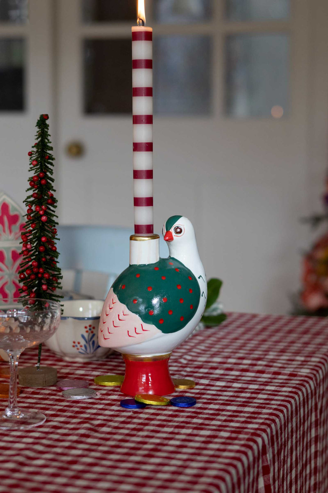 A decorative ceramic bird candle holder on a red-and-white checkered tablecloth, with a tall red-and-white striped candle inserted. The bird has a teal-green back with red polka dots, a white head and body, and a red base, with small scattered coins around it.