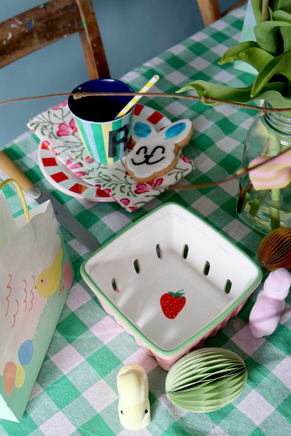 Table setting with a strawberry-shaped bowl, colorful mug, and decorative items on a checkered tablecloth.