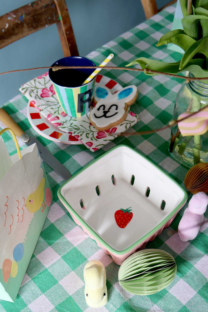 Table setting with a strawberry-shaped bowl, colorful mug, and decorative items on a checkered tablecloth.