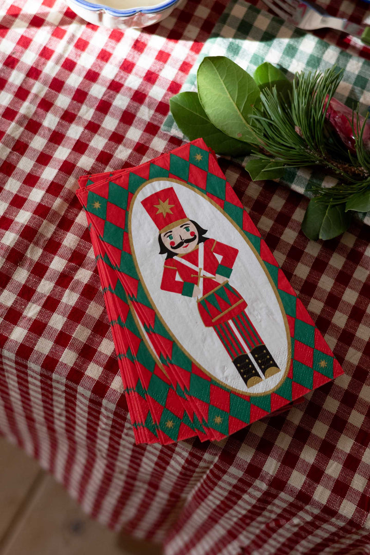 A stack of Christmas-themed place cards featuring a nutcracker illustration on a red and green checkered tablecloth with holiday greenery.