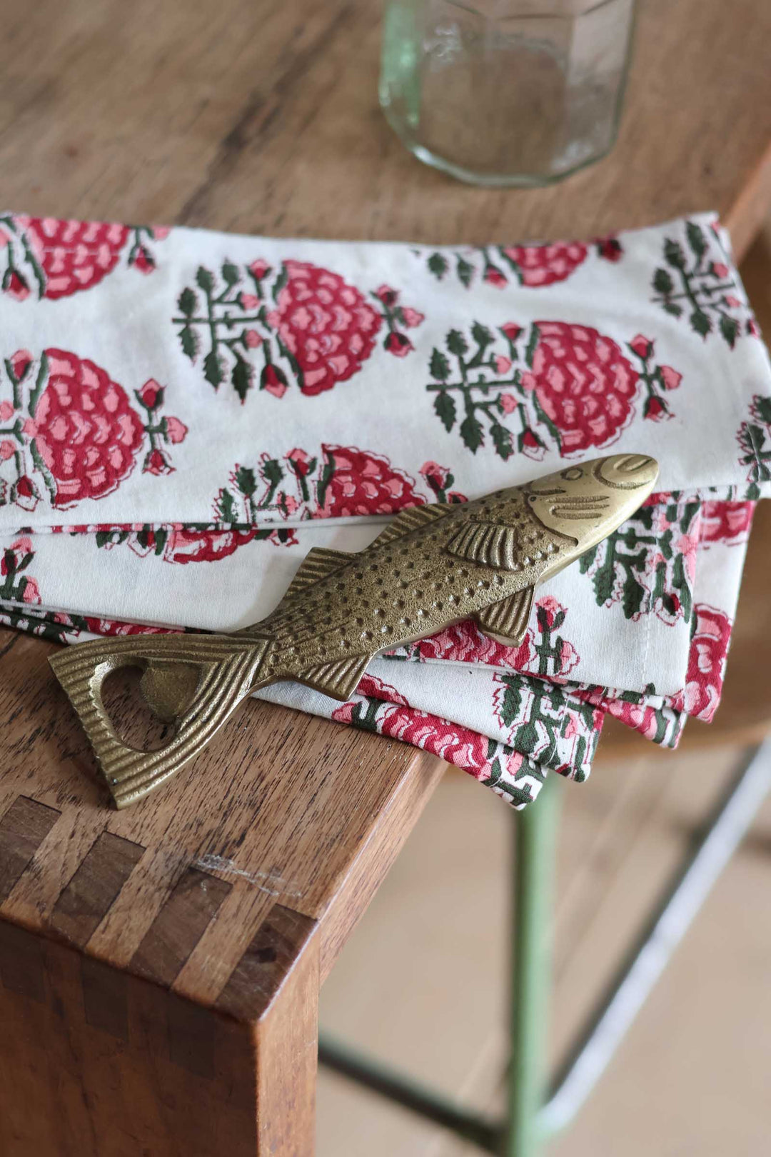 A folded white fabric napkin with a red and green floral block print pattern, styled on a wooden table with a brass fish-shaped bottle opener.
