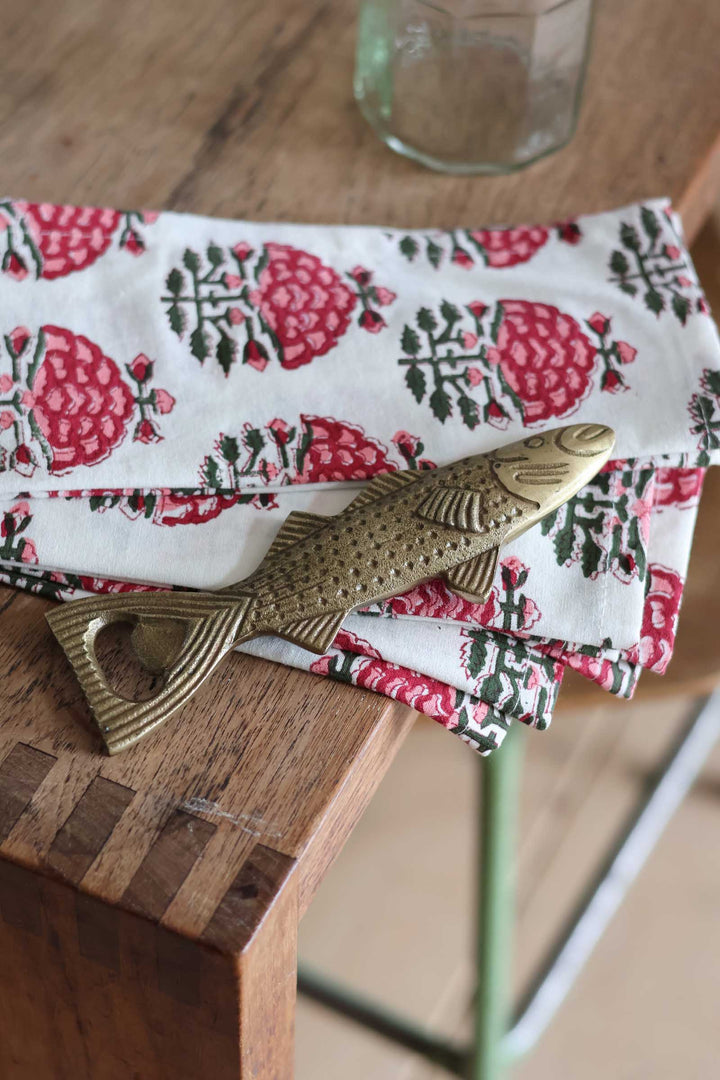A folded white fabric napkin with a red and green floral block print pattern, styled on a wooden table with a brass fish-shaped bottle opener.