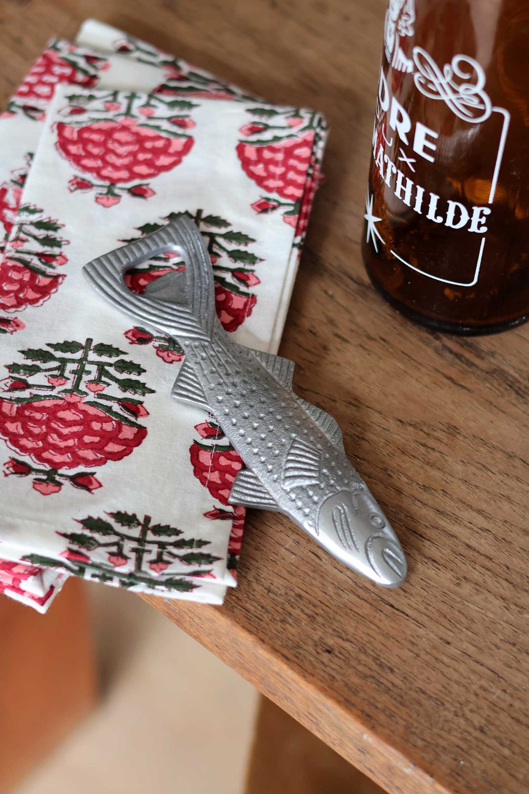 A silver metal fish-shaped bottle opener resting on a white napkin with a red and green pinecone pattern, placed on a wooden tabletop next to a brown glass bottle.