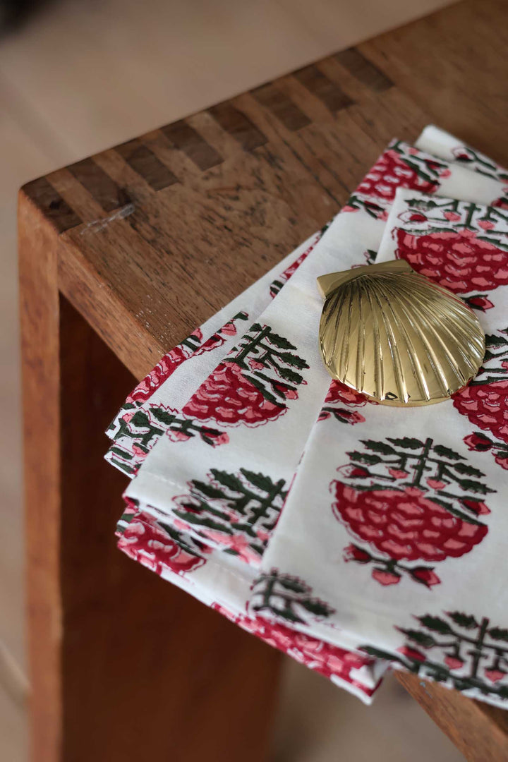 A stack of white napkins with a red and green botanical block print pattern, styled on a wooden table next to a gold scallop shell.